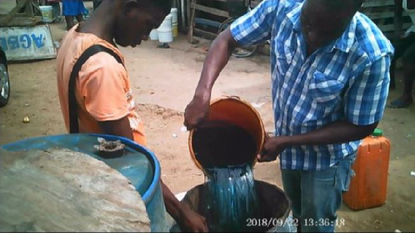 Two men pour a dark liquid from an orange bucket into a blue barrel in an outdoor workshop area.