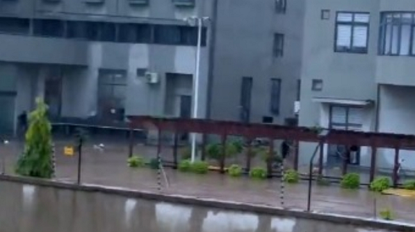 Flooded urban street beside gray apartment buildings; water covers the curb and a row of planters under a metal pergola.