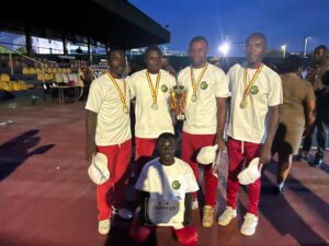 Five athletes in white shirts and red pants pose with medals and a trophy on a sports field; one kneels holding a certificate, stadium seating in background.