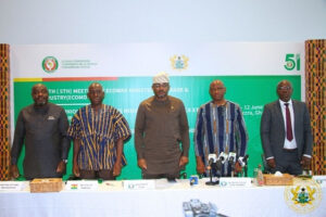 Five men stand behind a white conference table with microphones, during a formal press briefing; a green backdrop bearing official logos and text suggests an ECOWAS ministerial meeting, with one man wearing a traditional cap.