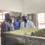 Group of men on a factory tour inspecting a can-sealing machine with rows of green cans.