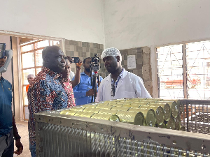 Group of men on a factory tour inspecting a can-sealing machine with rows of green cans.