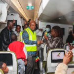 Inside a crowded airplane aisle, a man in a neon vest shakes hands with a passenger while others cheer and wave Ghana flags, capturing the welcome moment.