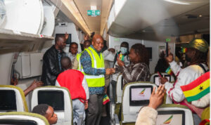Inside a crowded airplane aisle, a man in a neon vest shakes hands with a passenger while others cheer and wave Ghana flags, capturing the welcome moment.
