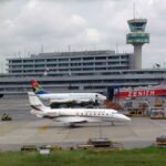 A small jet on the tarmac at an airport with a control tower and multi-level terminal in the background, a colorful tail fin visible on the aircraft.