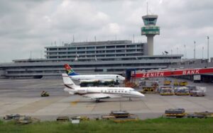 A small jet on the tarmac at an airport with a control tower and multi-level terminal in the background, a colorful tail fin visible on the aircraft.