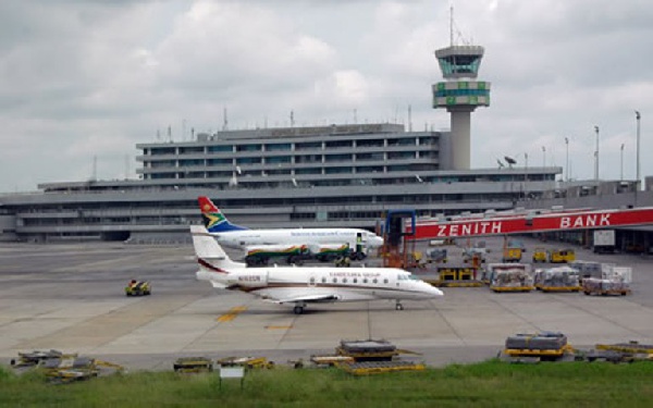 A small jet on the tarmac at an airport with a control tower and multi-level terminal in the background, a colorful tail fin visible on the aircraft.