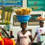 Three women carry baskets on their heads in a busy street market; one basket holds a pile of fries in the foreground.