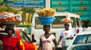 Three women carry baskets on their heads in a busy street market; one basket holds a pile of fries in the foreground.