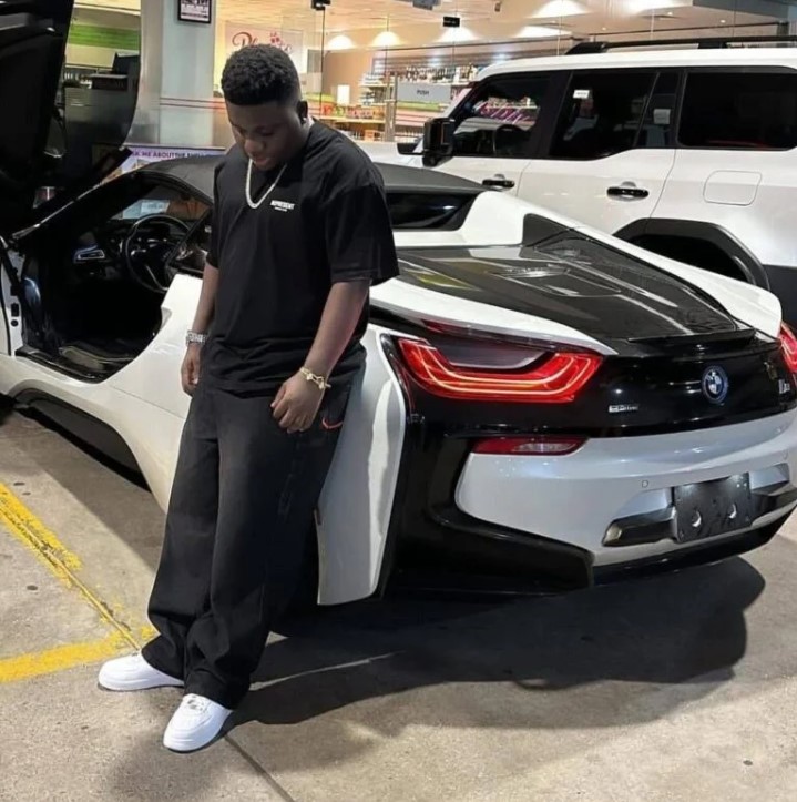 Young man in a black T-shirt and white sneakers stands beside a white sports car with its door open in a showroom.