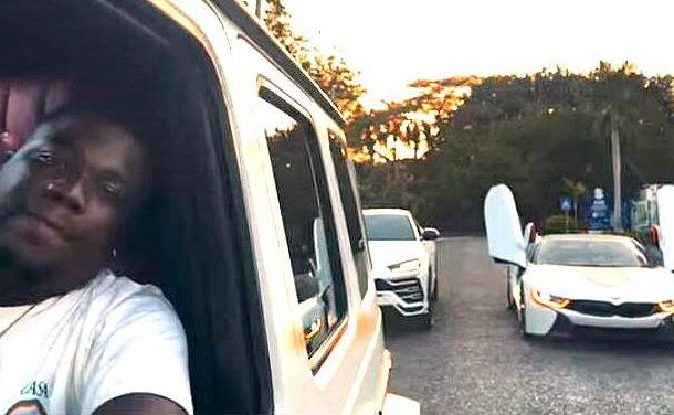 Person in a white shirt leaning from a white van window, taking a selfie, with two white cars with their gull-wing doors open in the background at sunset.