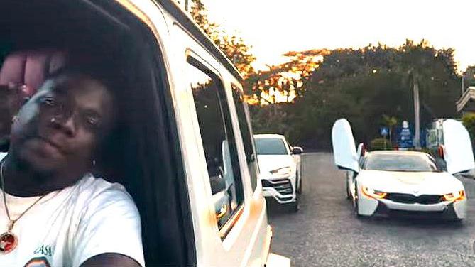Person in a white shirt leaning from a white van window, taking a selfie, with two white cars with their gull-wing doors open in the background at sunset.