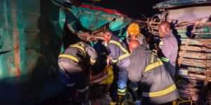 Nighttime rescue operation: firefighters and volunteers carry a person away from a crushed bus amid debris on a dark scene.