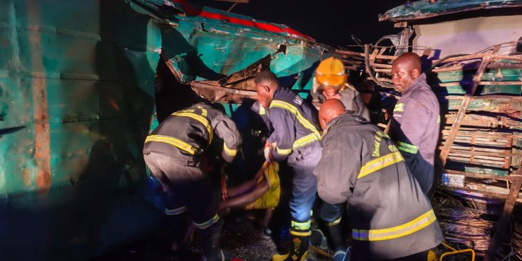Nighttime rescue operation: firefighters and volunteers carry a person away from a crushed bus amid debris on a dark scene.