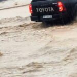 Rear view of a black Toyota pickup splashing through muddy floodwater, taillights glowing.