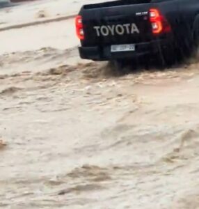 Rear view of a black Toyota pickup splashing through muddy floodwater, taillights glowing.