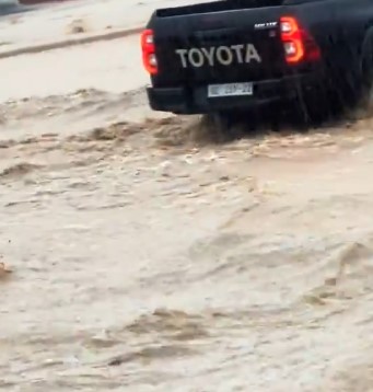 Rear view of a black Toyota pickup splashing through muddy floodwater, taillights glowing.