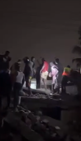 Nighttime scene of a crowd gathered on a rocky shoreline, with several people standing around a person on the ground wearing a red jacket and others in bright clothing.