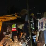 Nighttime rescue scene with workers clearing rubble from a collapsed building; a yellow crane arm visible in the left foreground.