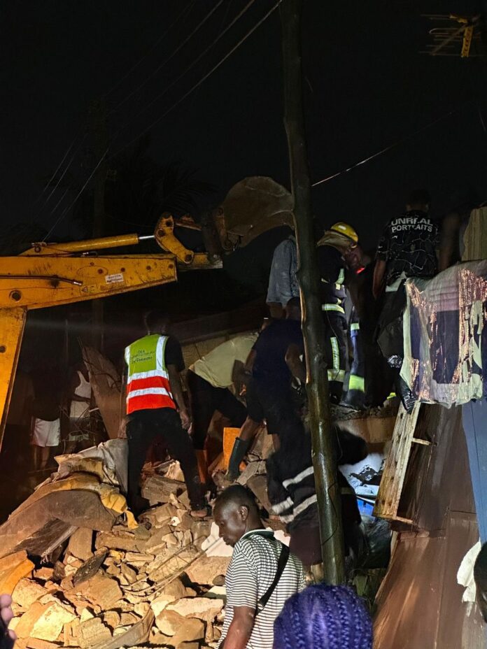 Nighttime rescue scene with workers clearing rubble from a collapsed building; a yellow crane arm visible in the left foreground.