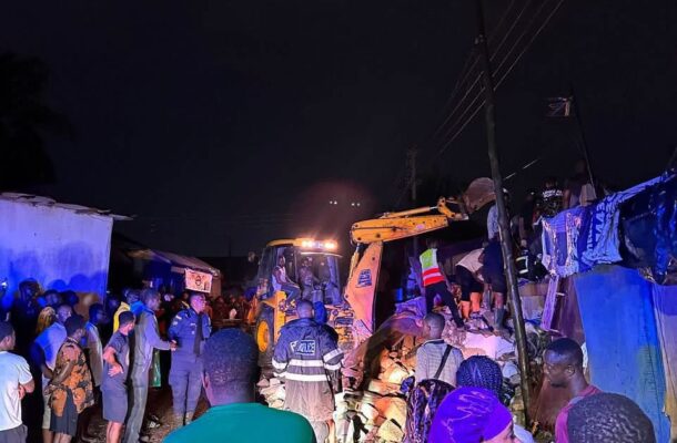 Nighttime rescue scene with a yellow excavator moving rubble while a crowd watches amid blue tarps and police responders in a crowded street at night.