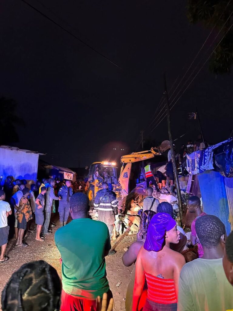 Nighttime rescue scene with a yellow excavator moving rubble while a crowd watches amid blue tarps and police responders in a crowded street at night.