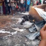A crowd of bystanders watches a charred, damaged object on the street; debris and ash are scattered on the ground, with a pixelated figure suggesting a person harmed in the incident.
