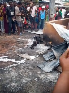 A crowd of bystanders watches a charred, damaged object on the street; debris and ash are scattered on the ground, with a pixelated figure suggesting a person harmed in the incident.