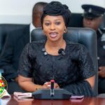 Ghana Parliament official woman in a black dress and headwrap sits at a desk with a microphone, with a Parliament Ghana seal and security officer in the background (informative).