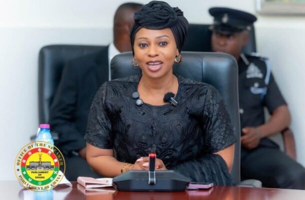 Adwoa Safo | Ghanaguardian.com Ghana Parliament official woman in a black dress and headwrap sits at a desk with a microphone, with a Parliament Ghana seal and security officer in the background (informative).