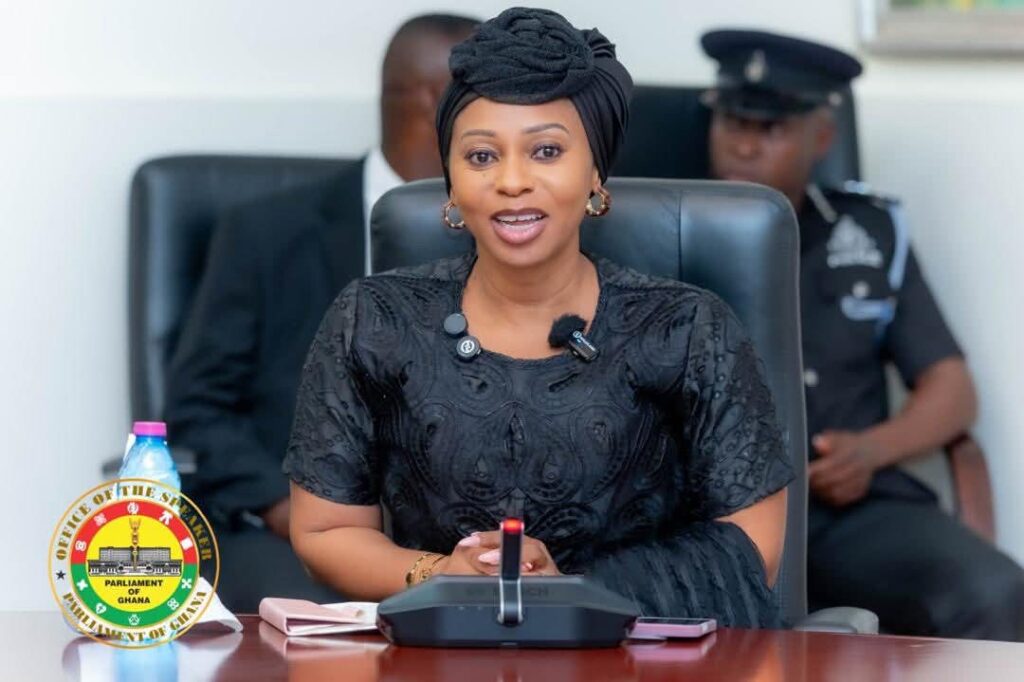 Adwoa Safo | Ghanaguardian.com Ghana Parliament official woman in a black dress and headwrap sits at a desk with a microphone, with a Parliament Ghana seal and security officer in the background (informative).