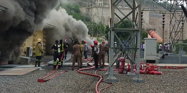 Firefighters battle a blaze with hoses as thick black smoke billows from a building area.