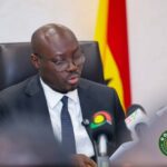 Man in a dark suit and glasses speaks at a press conference, with microphones in front and a Ghana flag in the background.