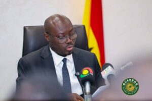 Man in a dark suit and glasses speaks at a press conference, with microphones in front and a Ghana flag in the background.