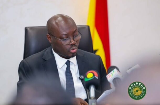 Man in a dark suit and glasses speaks at a press conference, with microphones in front and a Ghana flag in the background.