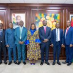 Nine professionals in formal attire pose for a group photo in a wood-paneled government office; woman in a blue and gold patterned dress stands center.