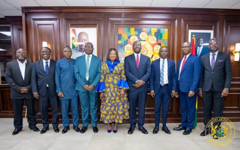 Nine professionals in formal attire pose for a group photo in a wood-paneled government office; woman in a blue and gold patterned dress stands center.