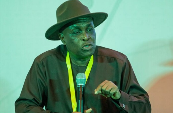Man wearing a wide-brimmed hat and dark traditional outfit speaks into a handheld microphone on stage, with a yellow lanyard around his neck.