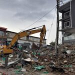 Yellow CAT hydraulic excavator amid a pile of rubble and debris in a demolished urban site, with a damaged multi-story building in the background.