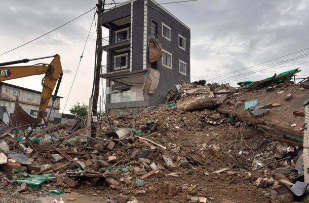Excavator amid a collapsed three-story building and surrounding construction rubble on a muddy lot under a cloudy sky, urban debris scattered everywhere.