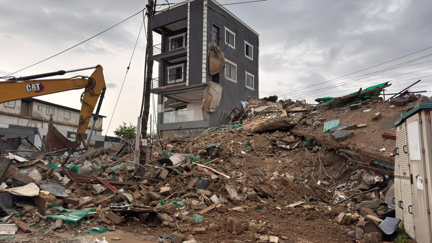 Excavator amid a collapsed three-story building and surrounding construction rubble on a muddy lot under a cloudy sky, urban debris scattered everywhere.