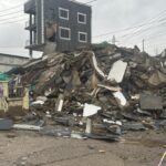 Rubble and debris pile in front of a damaged multi-story building after a collapse, with exposed rebar and broken walls under a cloudy sky