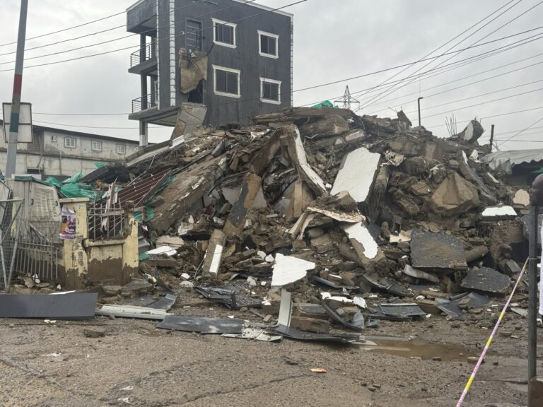 Rubble and debris pile in front of a damaged multi-story building after a collapse, with exposed rebar and broken walls under a cloudy sky