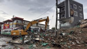 Yellow CAT hydraulic excavator amid a pile of rubble and debris in a demolished urban site, with a damaged multi-story building in the background.