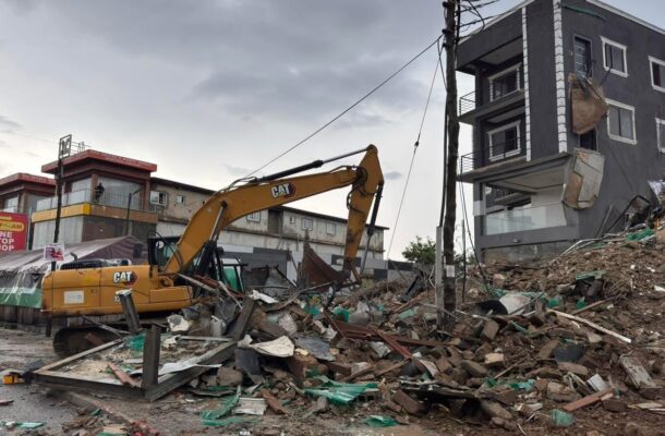 Avenor storey building collapse | Ghanaguardian.com Yellow CAT hydraulic excavator amid a pile of rubble and debris in a demolished urban site, with a damaged multi-story building in the background.