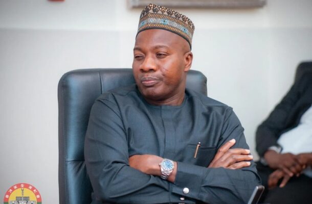 Man in a traditional cap and dark outfit sits with arms crossed in a padded office chair, looking contemplative.