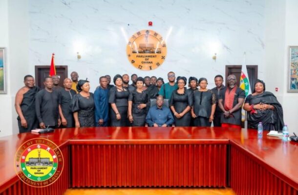 Bagbin 1 | Ghanaguardian.com Group of men and women in black formal attire posing for a group photo in the Parliament of Ghana chamber, with the emblem on the wall and flags visible.