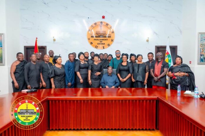 Group of men and women in black formal attire posing for a group photo in the Parliament of Ghana chamber, with the emblem on the wall and flags visible.