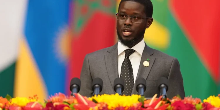 Man in a gray suit giving a speech at a podium with five microphones, colorful flags behind him.