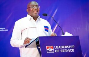 Man in a white shirt delivering a speech at a lectern with a blue banner reading 'Leadership of Service' on stage with a microphone nearby